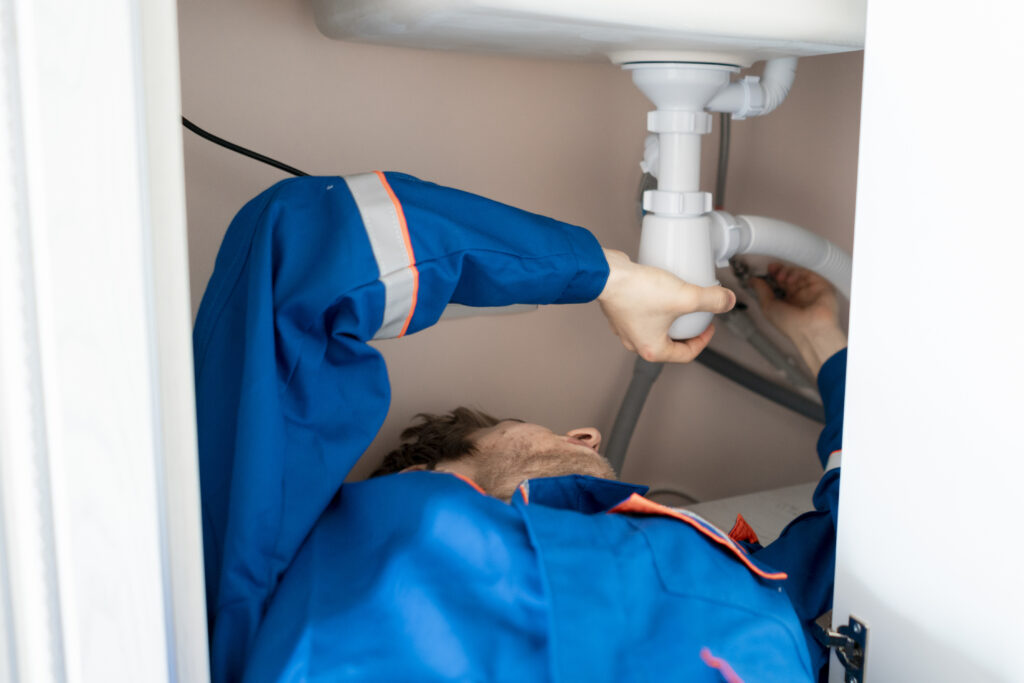 plumber in blue uniform lying in the kitchen repairing a leak